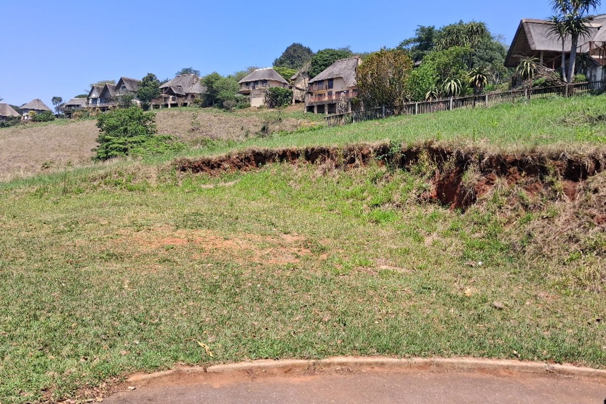 A vacant residential plot featuring a sloped grassy landscape with visible soil erosion, situated in a developed estate with surrounding homes.
