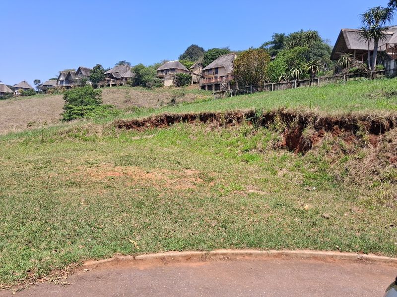 A vacant residential plot featuring a sloped grassy landscape with visible soil erosion, situated in a developed estate with surrounding homes.