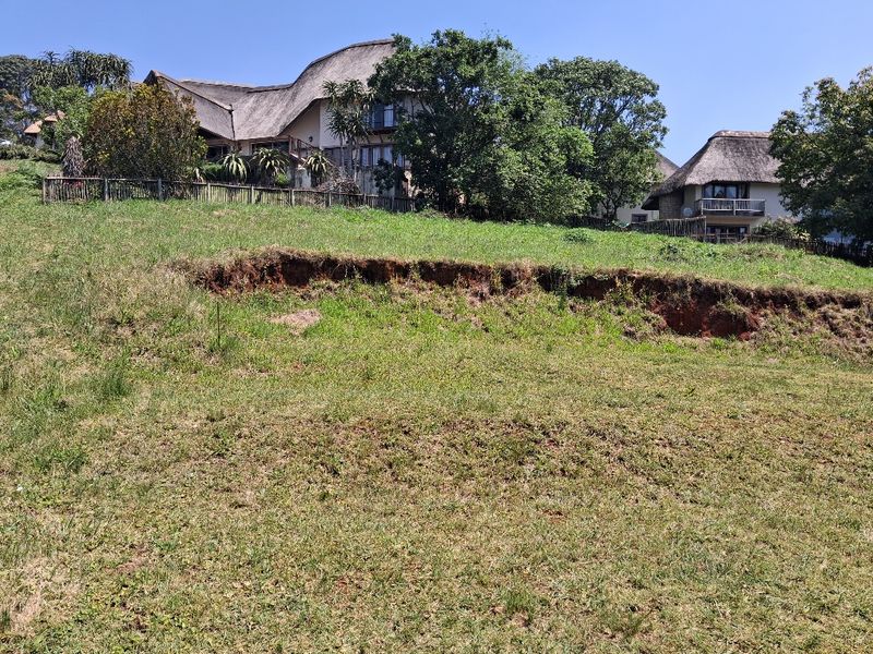 A sloped residential vacant land plot featuring natural grass cover and visible soil erosion, with established thatched-roof properties visible in the background.