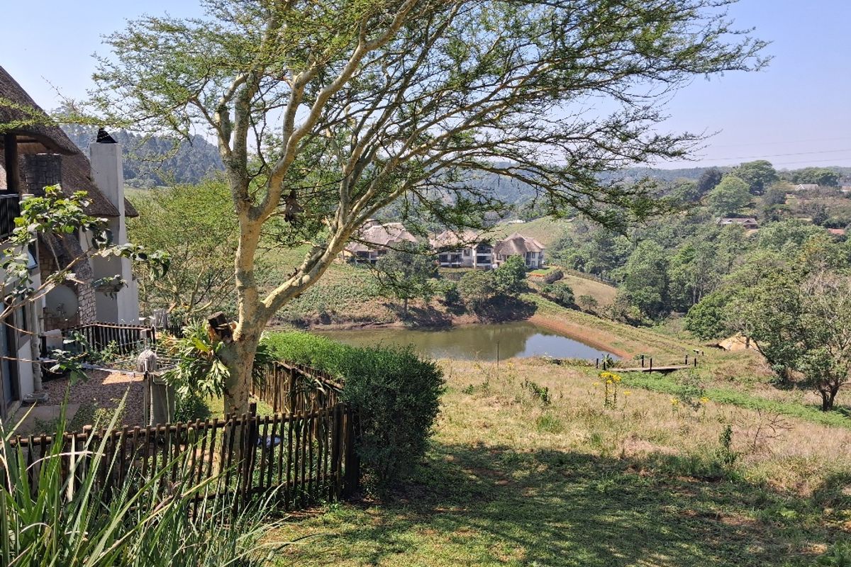 Scenic view overlooking a dam and surrounding landscape, featuring a portion of a thatched-roof residence with a stone chimney and wooden fencing.