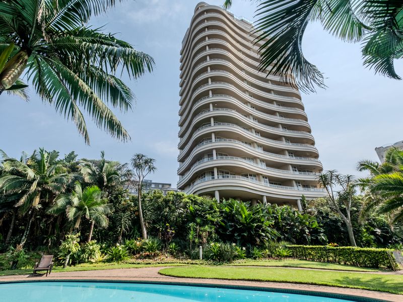 A view of a modern high-rise apartment building featuring curved balconies, set behind a well-maintained swimming pool and lush landscaped garden area.