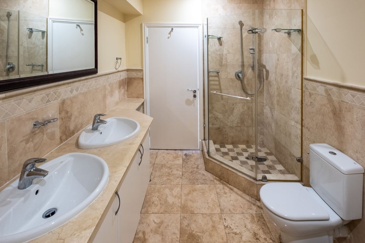 A well-maintained bathroom featuring a double vanity with stone countertop, glass-enclosed shower, and neutral stone tiling throughout.