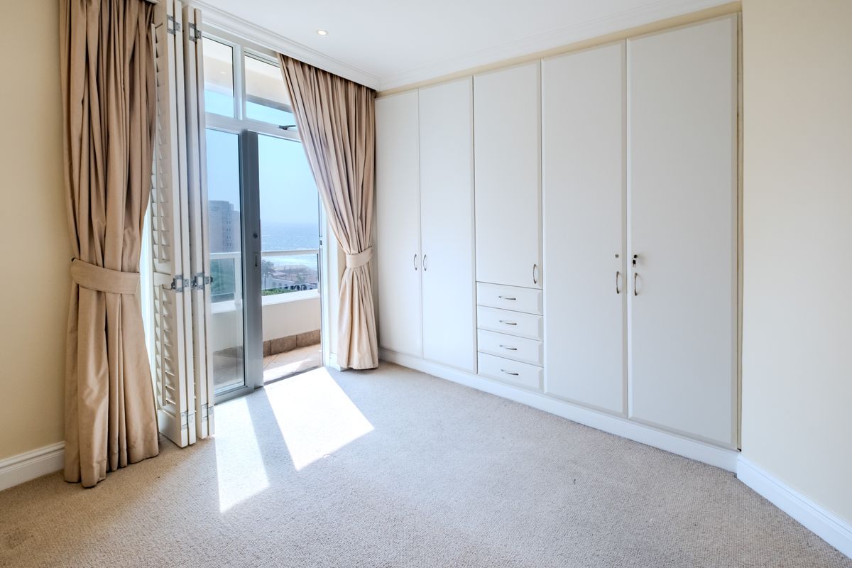 A bright, empty bedroom featuring extensive built-in cupboards, neutral carpeting, and a sliding glass door opening onto a balcony with ocean views.