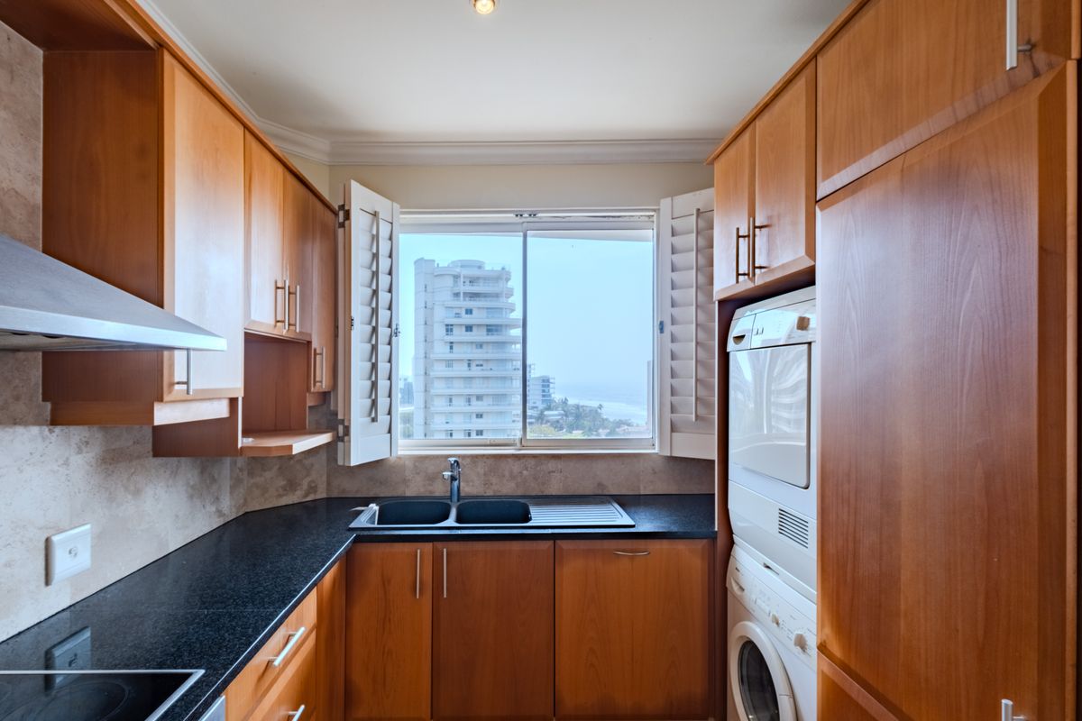 Compact kitchen featuring warm wooden cabinetry, granite countertops, a double sink, and integrated laundry appliances with a view of the surrounding buildings.