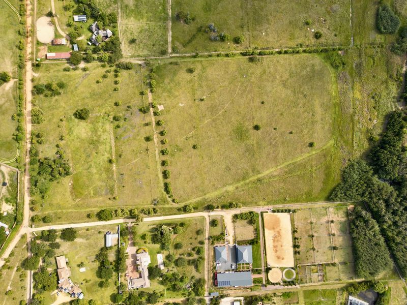 Aerial view of a large residential vacant land plot featuring open pasture, fenced paddocks, a riding arena, and established trees.
