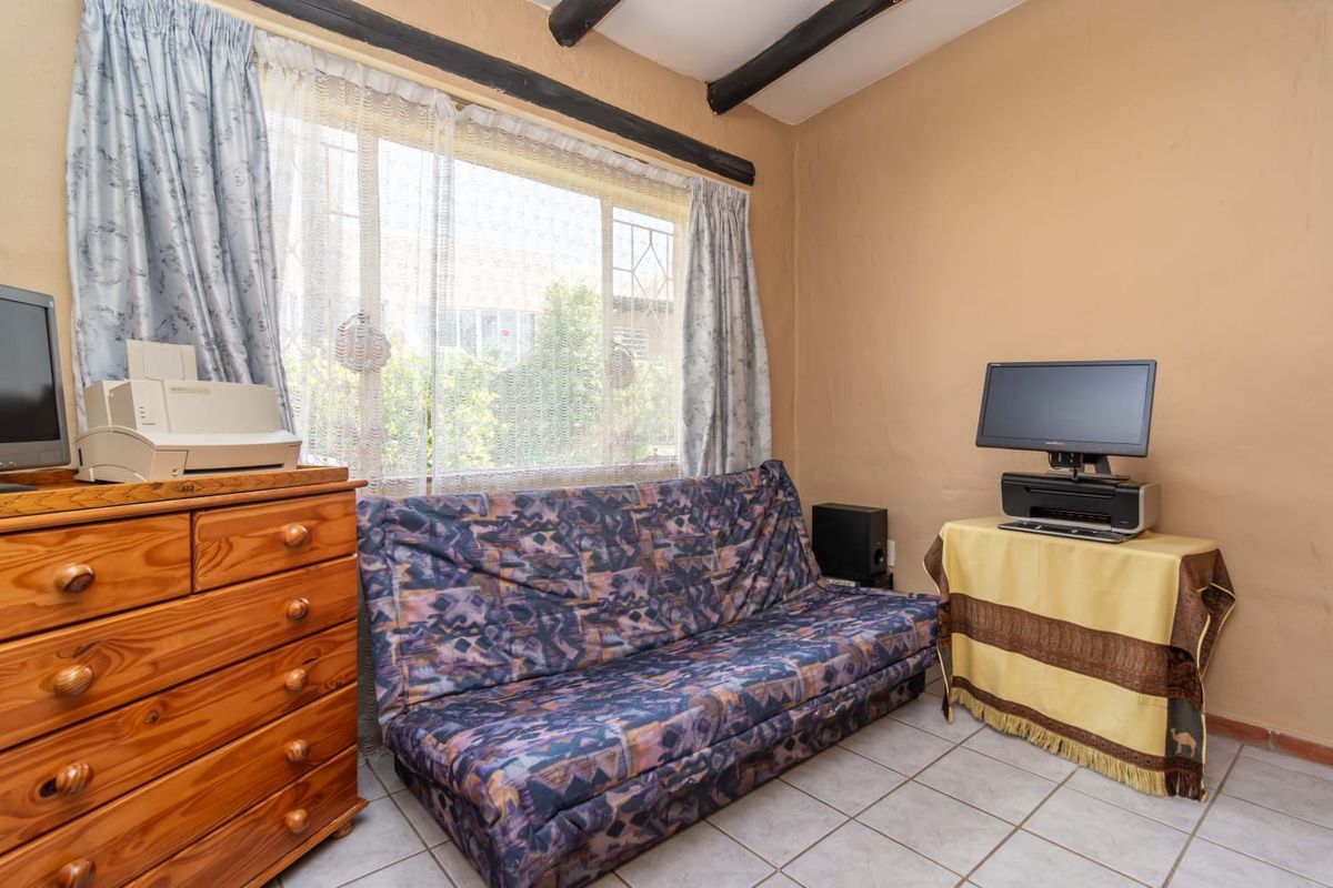 A compact bedroom featuring tiled flooring, exposed ceiling beams, and a large window with security bars, offering a functional space with potential for modernization.