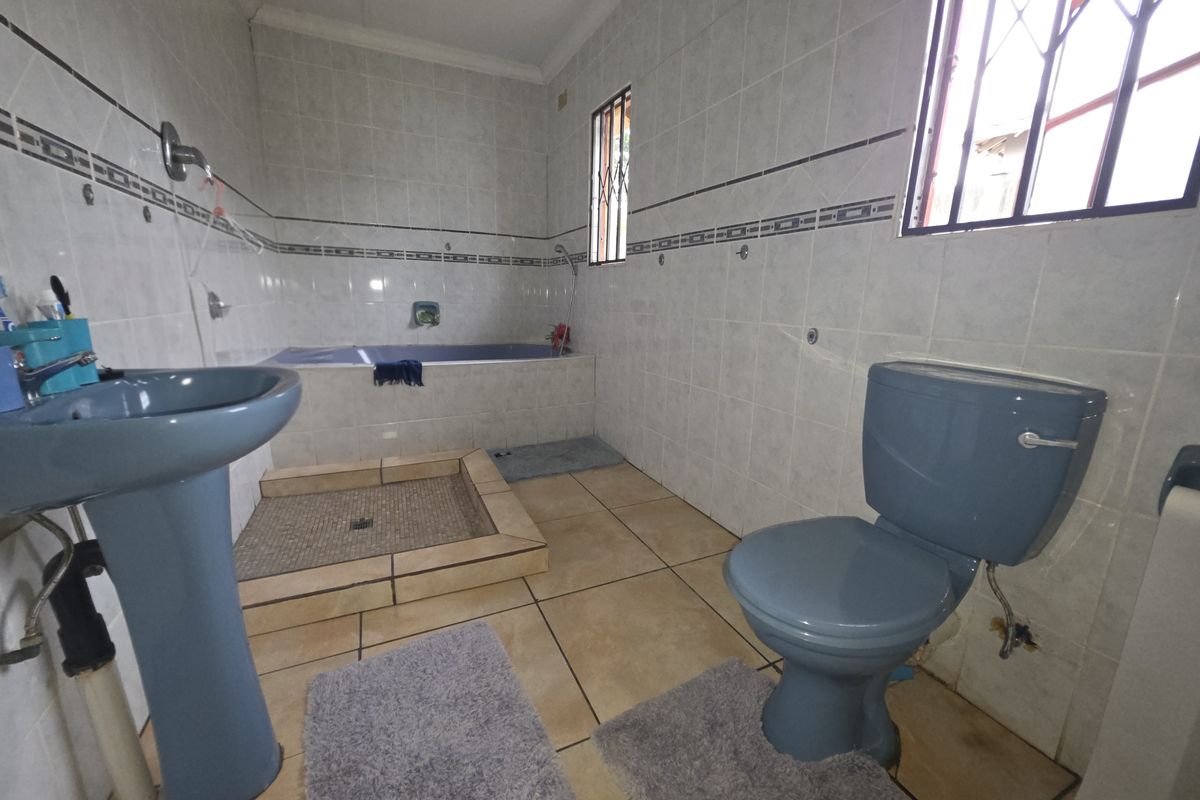 A dated bathroom featuring a blue pedestal basin, matching toilet, built-in bathtub, and a tiled shower base with security bars on the windows.