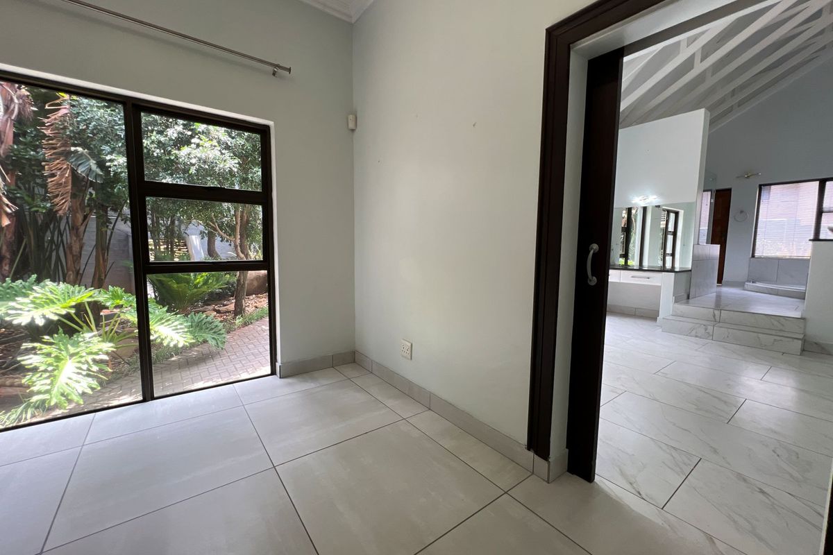 A bright, empty room featuring large windows with garden views, tiled flooring, and a sliding door leading to an adjacent bathroom area.