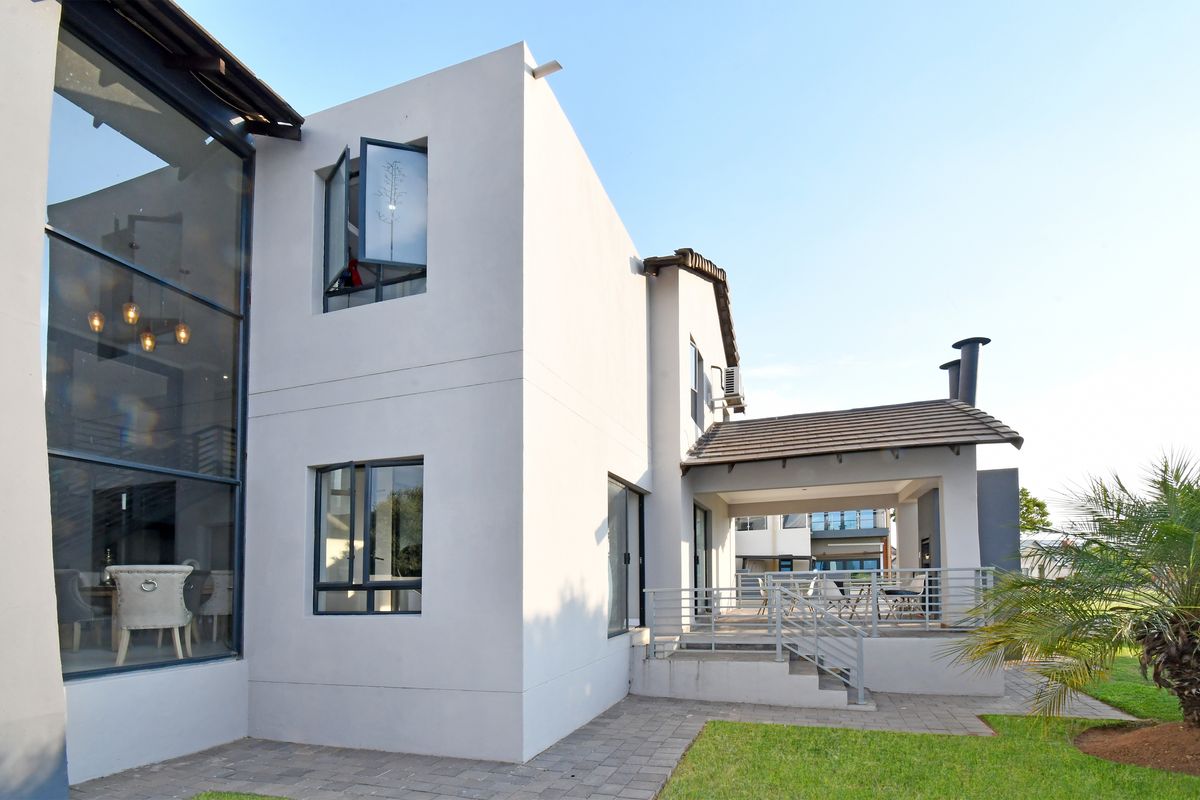 Rear view of a modern double-story home featuring a double-volume glass facade, a covered patio with an external fireplace, and a landscaped garden.
