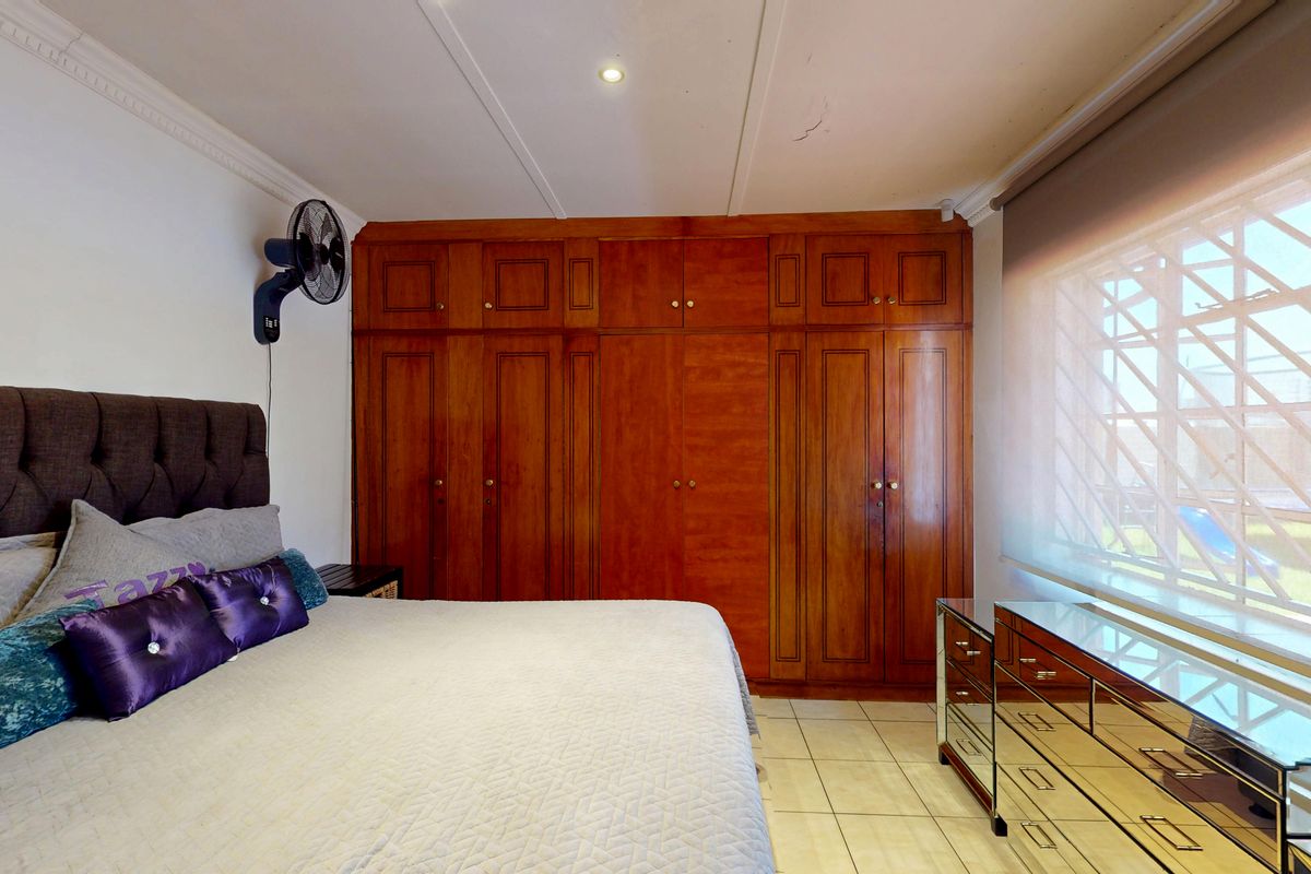 A bedroom featuring extensive built-in wooden cabinetry, tiled flooring, and a large window with security bars and a roller blind.