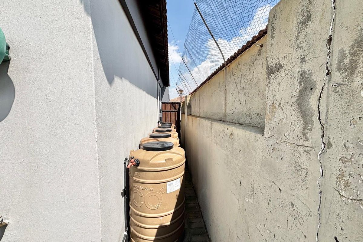 A narrow side passage featuring a row of water storage tanks installed against the townhouse exterior wall and a weathered boundary wall with security fencing.