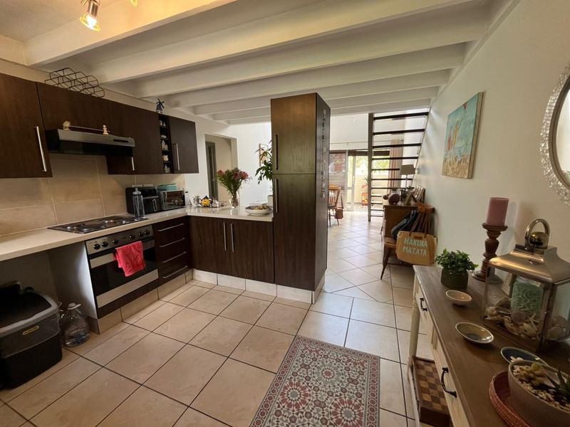 Open-plan kitchen featuring dark wood cabinetry, built-in oven and hob, tiled flooring, and exposed ceiling beams, leading towards a staircase and living area.