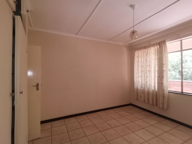 A bright, empty bedroom featuring neutral tiled flooring, a built-in cupboard, and a large window with natural light.