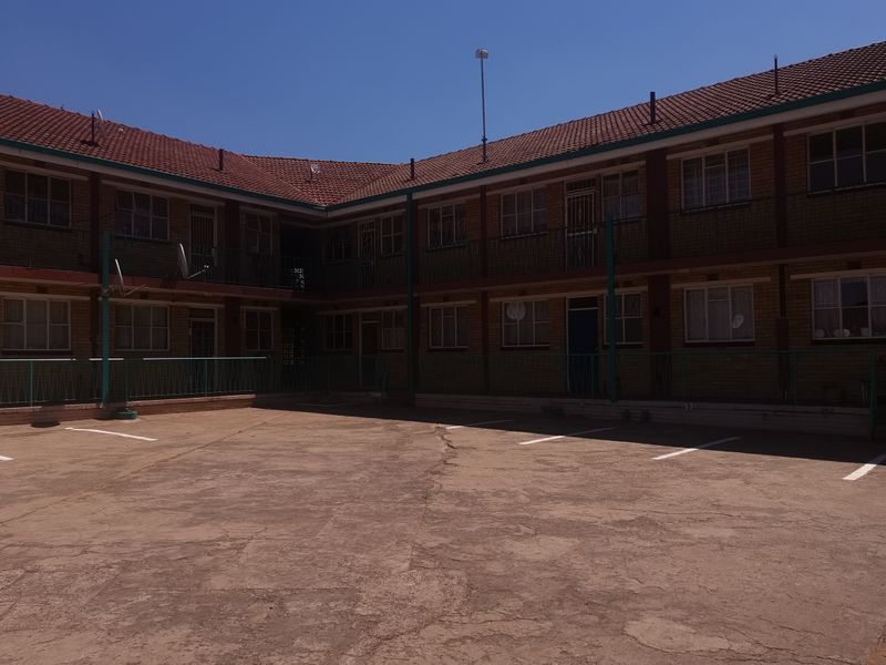 Exterior view of a multi-story brick apartment complex featuring a paved courtyard with designated parking bays and metal balcony railings.