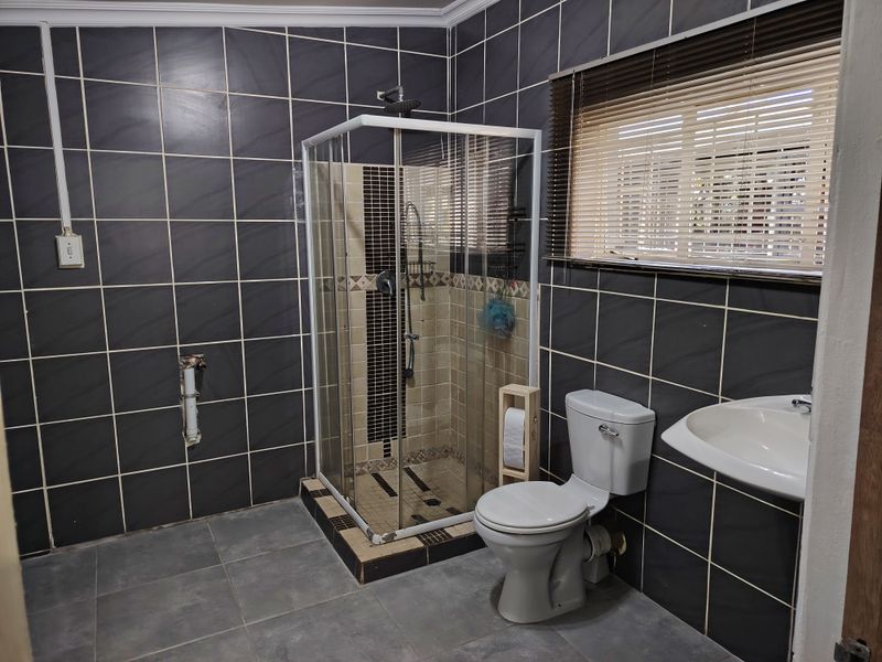 A functional bathroom featuring dark wall tiles, a corner shower enclosure, a standard toilet, and a wall-mounted washbasin with natural light from the window.