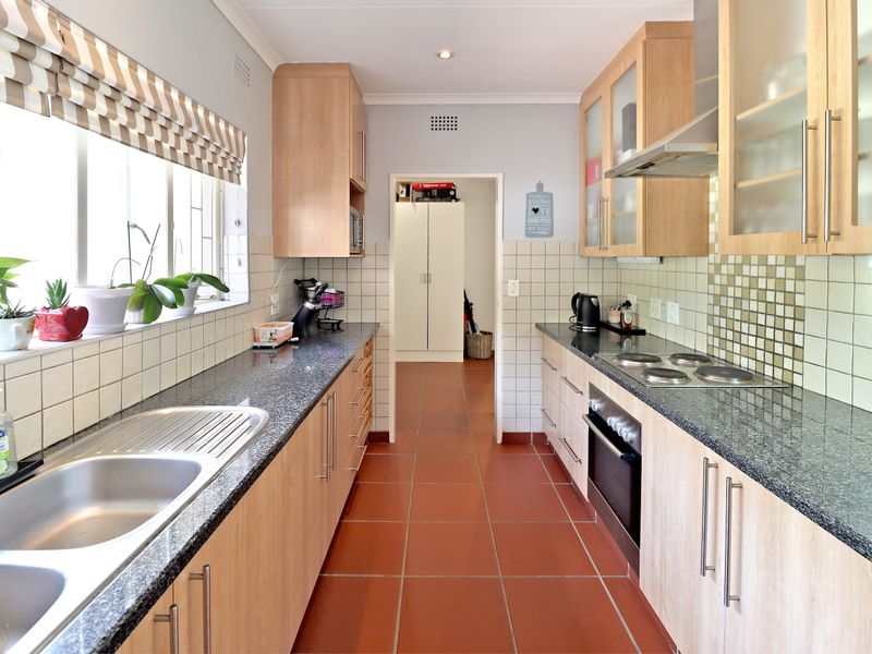 A functional galley-style kitchen featuring light wood cabinetry, granite countertops, a built-in oven, electric hob, and tiled flooring.