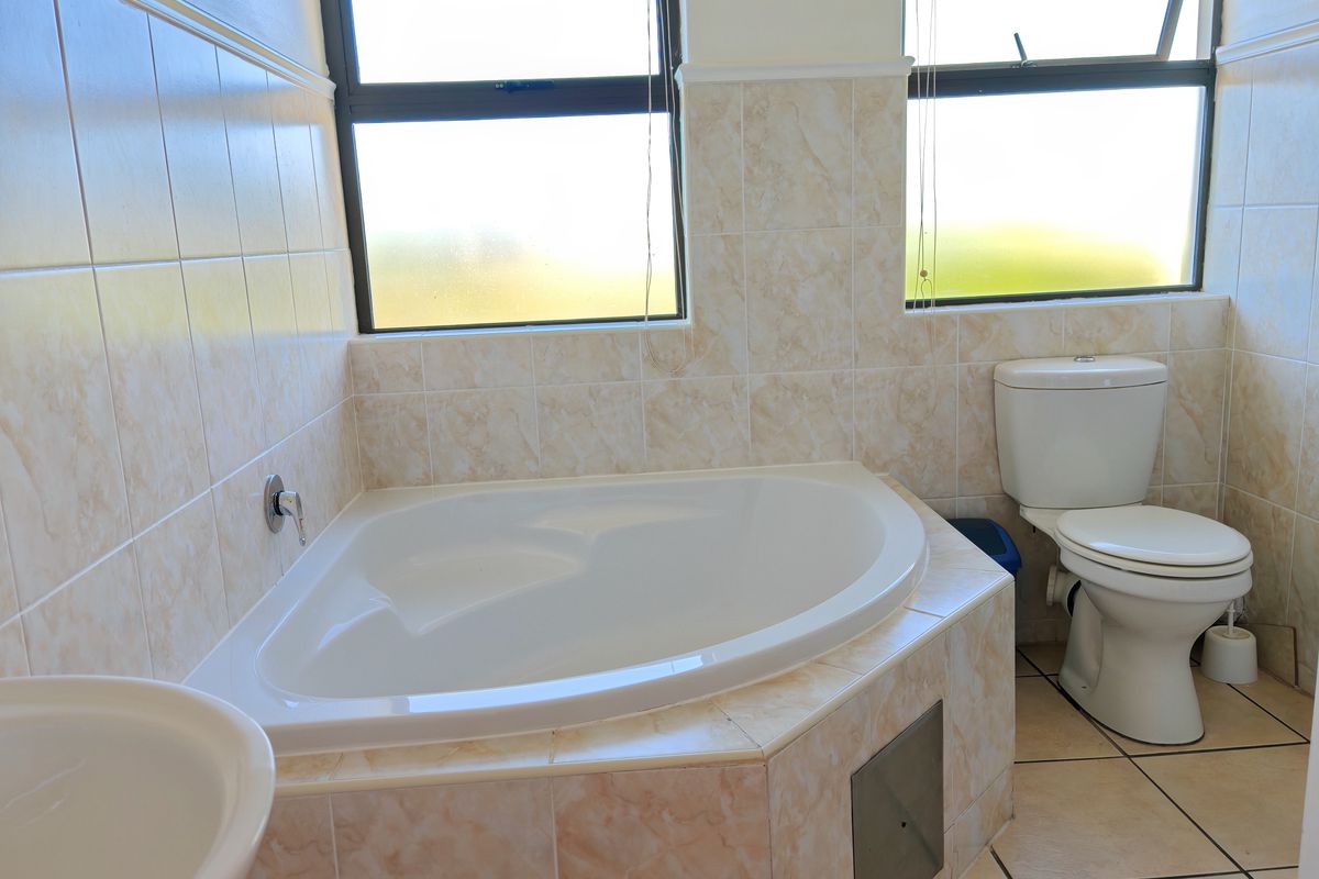 A functional bathroom featuring a corner bathtub, toilet, and neutral tiled finishes with natural light from two windows.