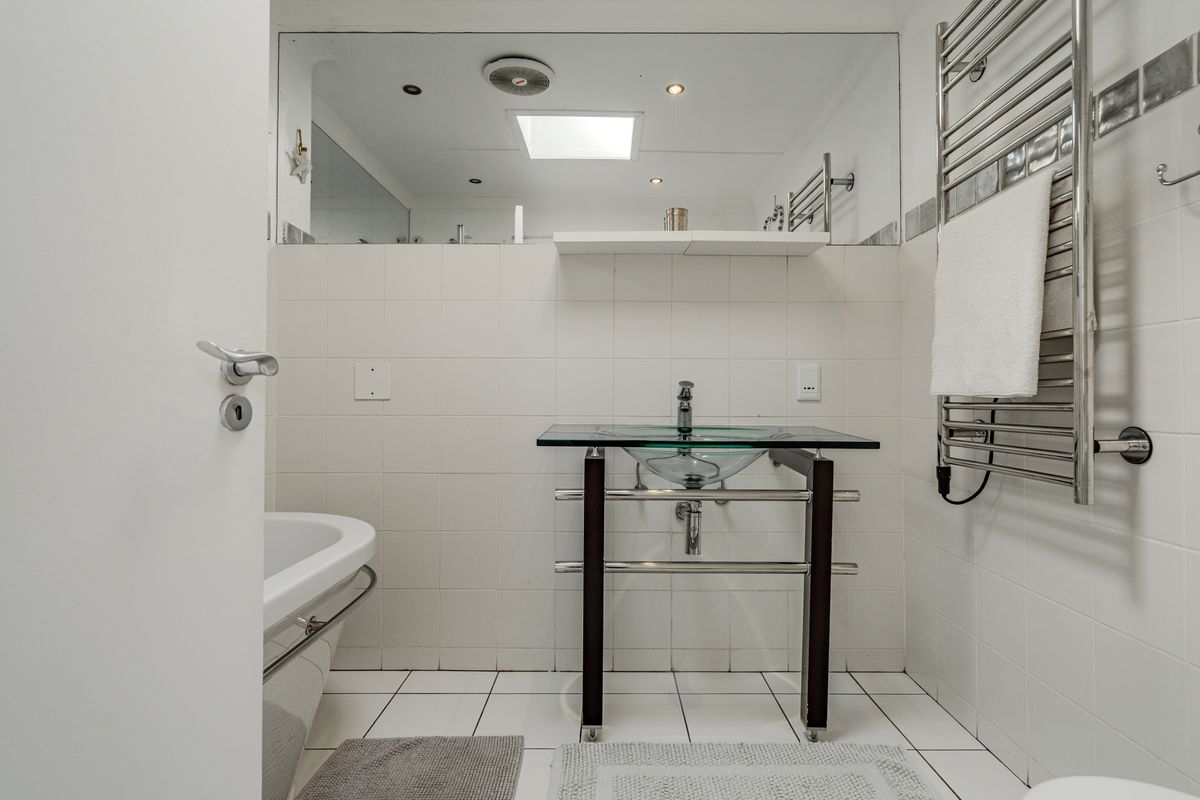 A bright bathroom featuring a modern glass vessel sink, chrome heated towel rail, and a skylight, finished with clean white wall and floor tiling.