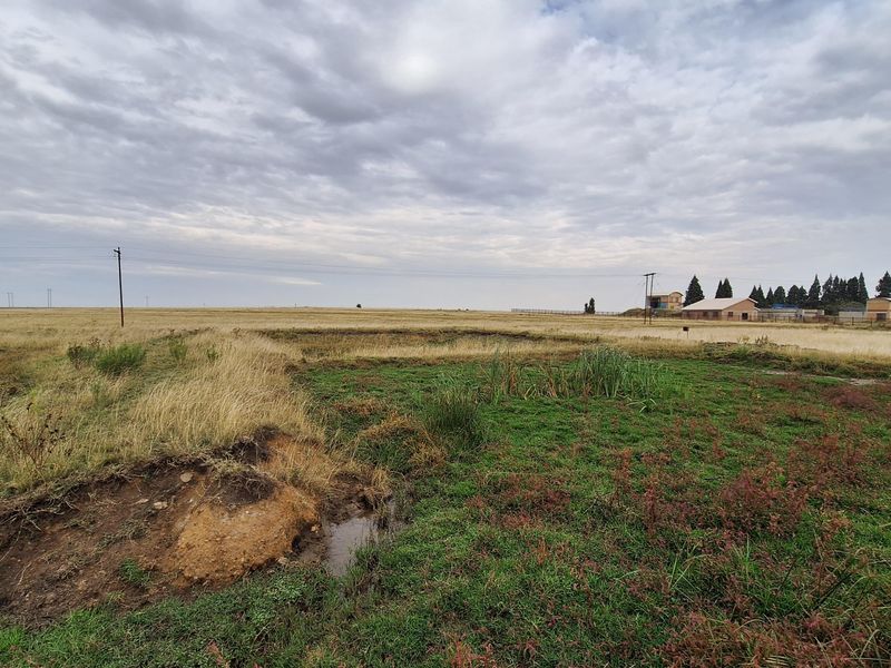 A wide-angle view of a vacant residential plot featuring natural grassland, uneven terrain, and visible utility infrastructure under a cloudy sky.