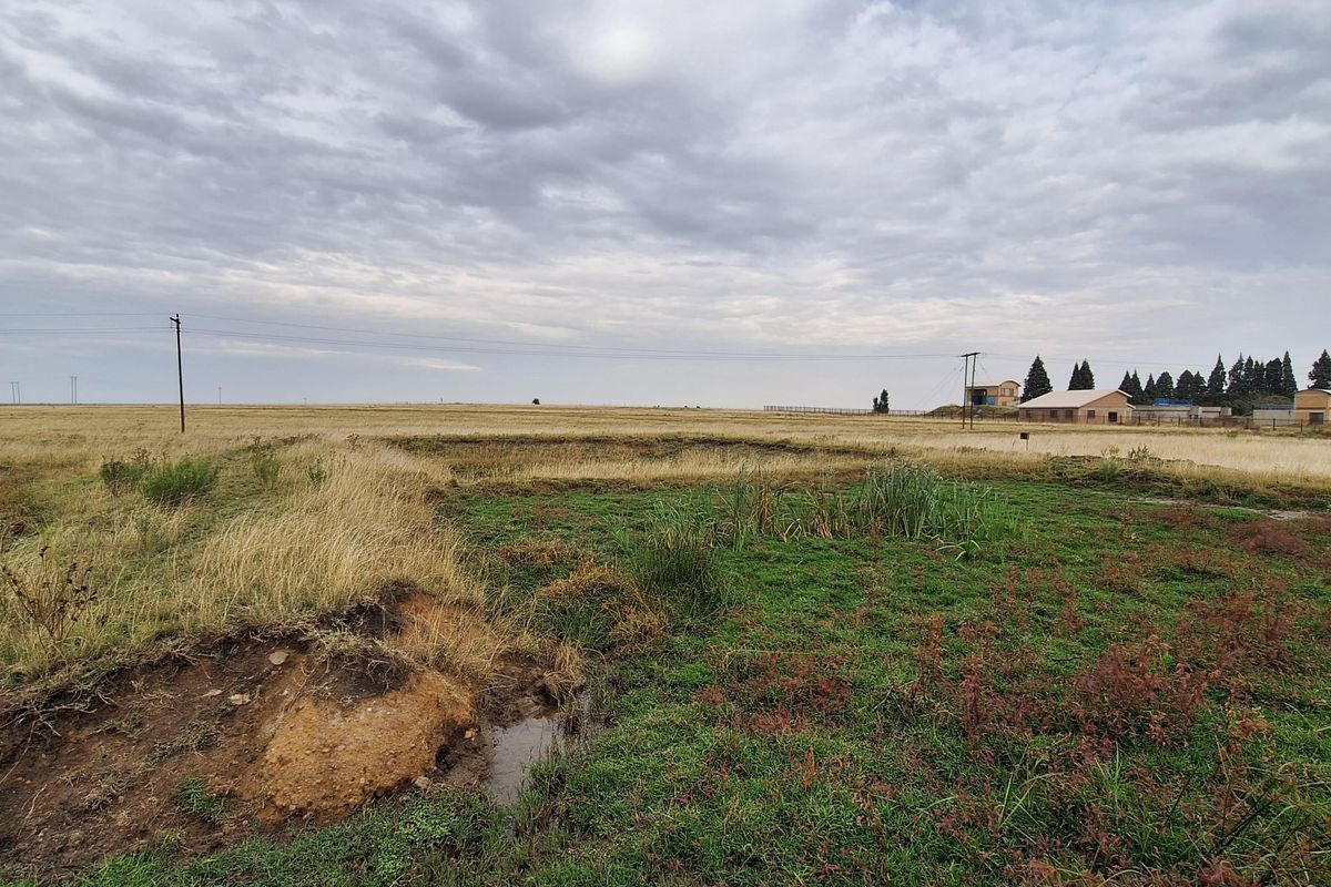 A wide-angle view of a vacant residential plot featuring natural grassland, uneven terrain, and visible utility infrastructure under a cloudy sky.
