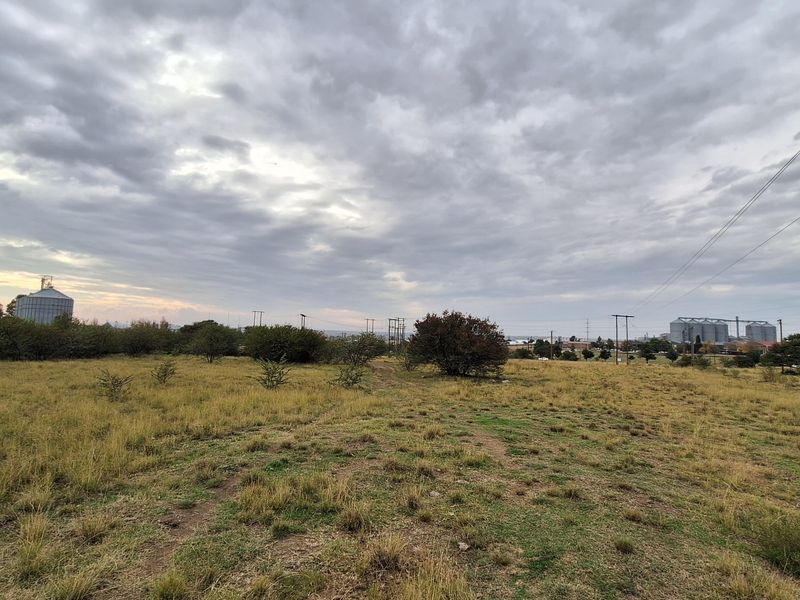 A wide-angle view of a vacant residential plot featuring natural grassland and distant industrial structures under a cloudy sky.