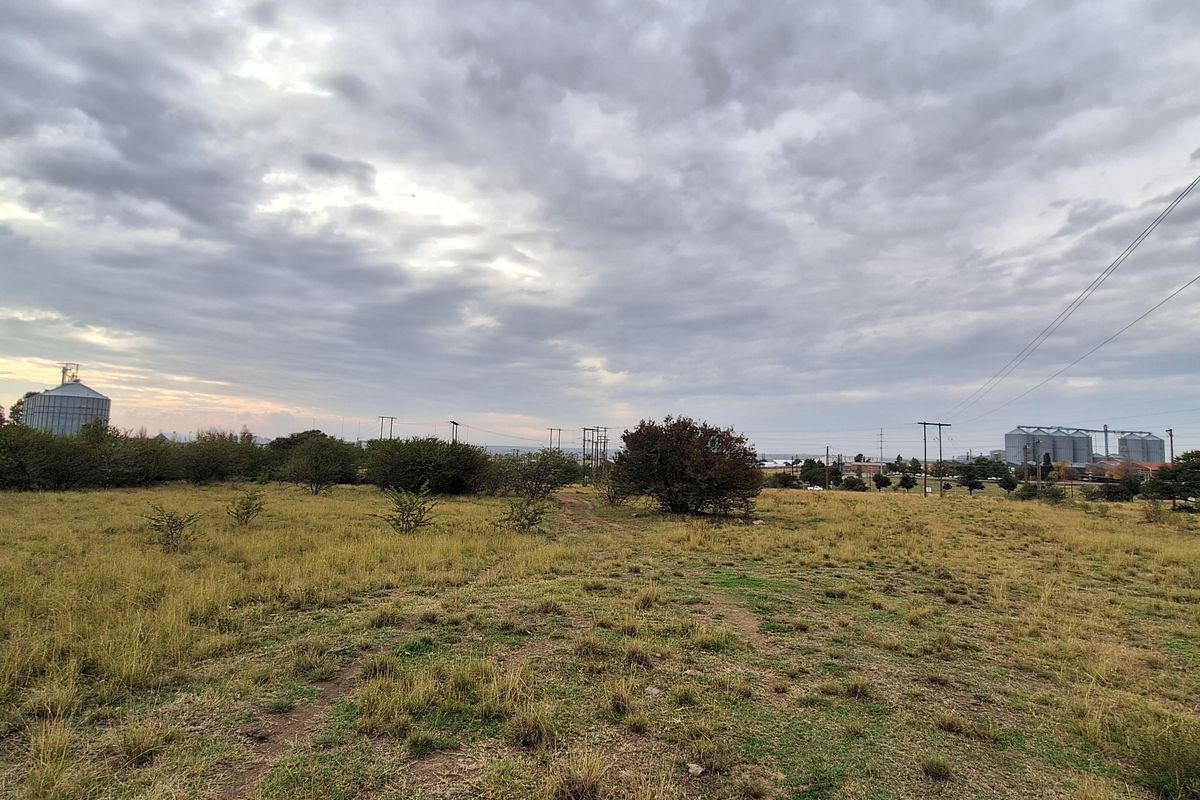 A wide-angle view of a vacant residential plot featuring natural grassland and distant industrial structures under a cloudy sky.