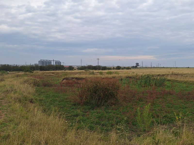 An expansive, undeveloped residential plot featuring natural grassland and distant industrial structures under an overcast sky.