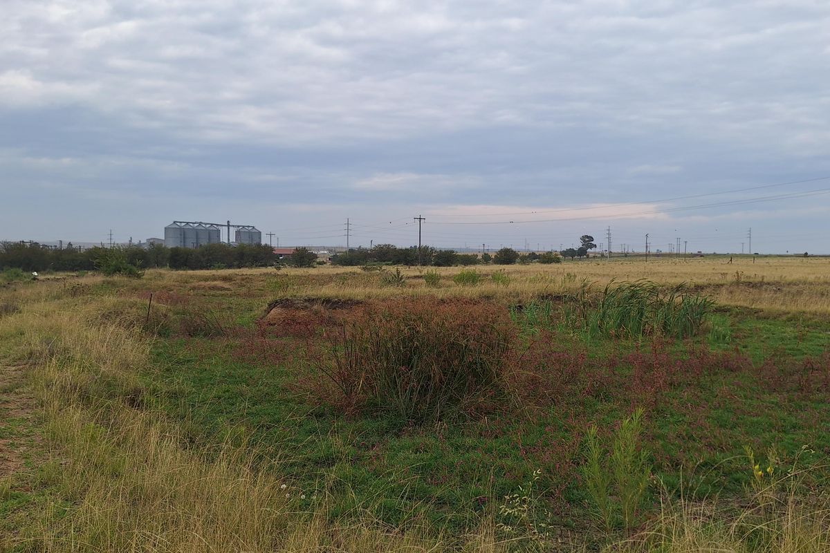 An expansive, undeveloped residential plot featuring natural grassland and distant industrial structures under an overcast sky.