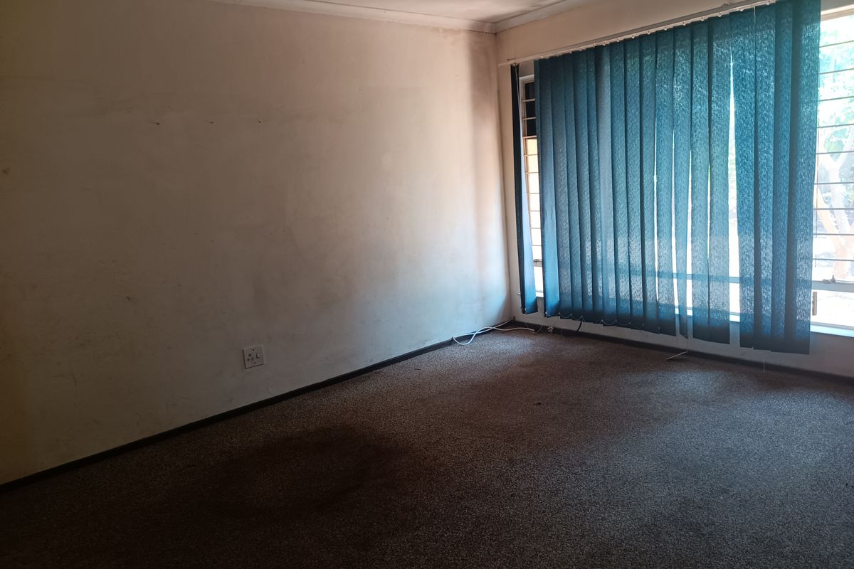 An empty living room featuring wall-to-wall carpeting and large windows with vertical blinds, requiring cosmetic updates to improve the overall aesthetic.