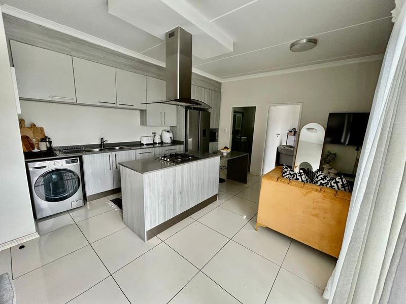 Modern open-plan kitchen and living area featuring light wood-grain cabinetry, a central island with gas hob, and sleek tiled flooring throughout.
