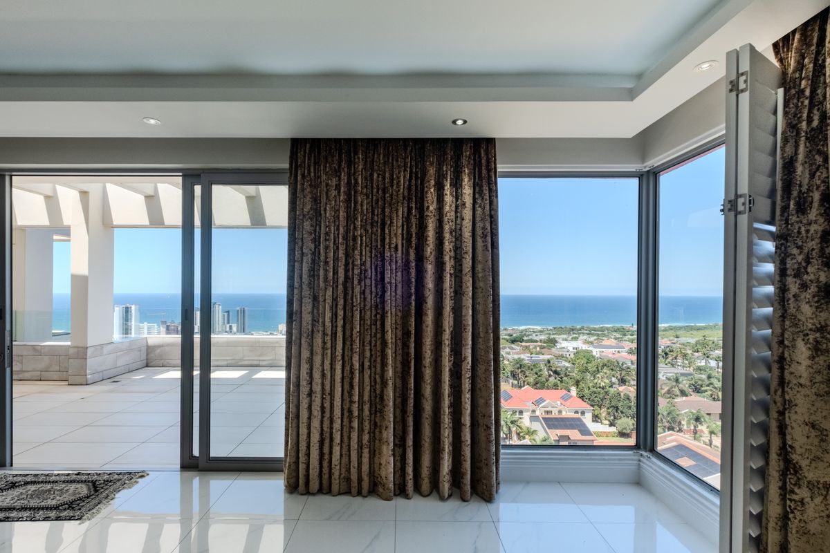 Spacious living area featuring large windows and sliding glass doors that open onto a wide balcony with expansive ocean and coastal views.