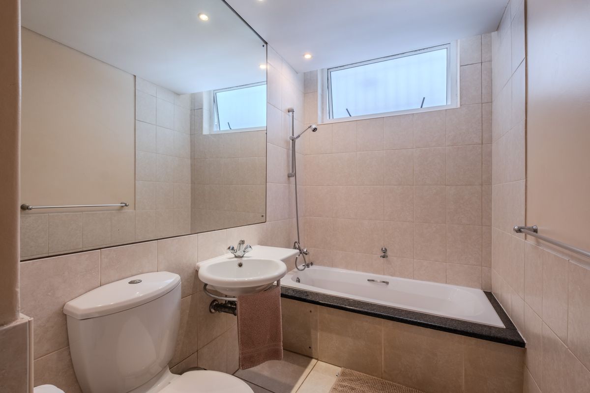 A functional bathroom featuring a built-in bathtub with shower attachment, pedestal basin, toilet, and large wall mirror with neutral wall and floor tiling.