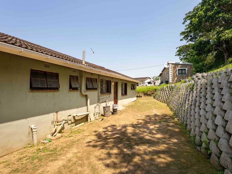 Rear exterior view of the property featuring a concrete block retaining wall, tiled roof, and exposed exterior plumbing along the side facade.
