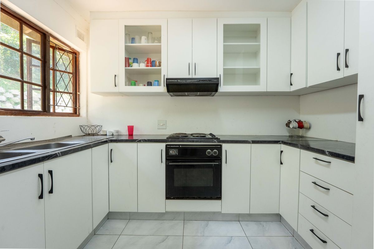 A functional kitchen featuring white cabinetry, dark laminate countertops, built-in oven with hob, extractor fan, and tiled flooring with natural light from a side window.