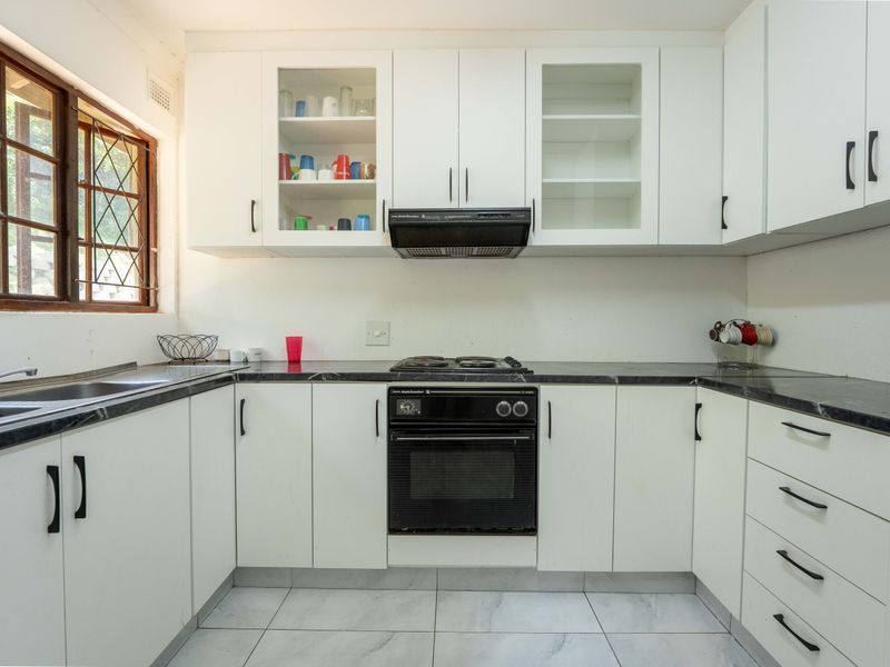 A functional kitchen featuring white cabinetry, dark laminate countertops, built-in oven with hob, extractor fan, and tiled flooring with natural light from a side window.
