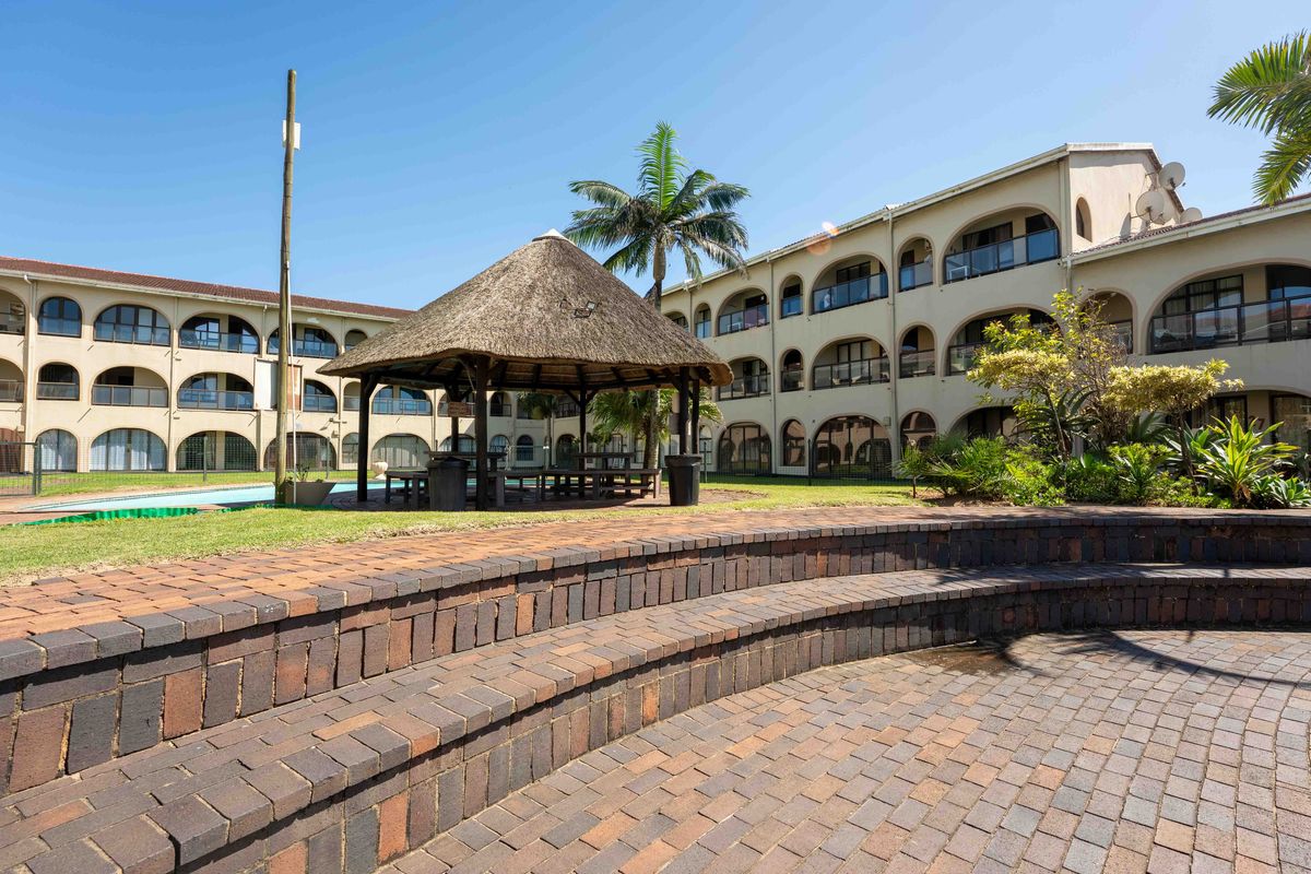 A communal outdoor area featuring a thatched gazebo, paved seating steps, and landscaped gardens set within an apartment complex.