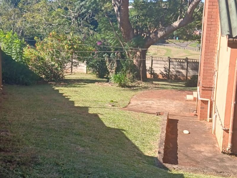 A rear garden view featuring a brick building facade, a paved patio area, and a spacious lawn with mature trees.