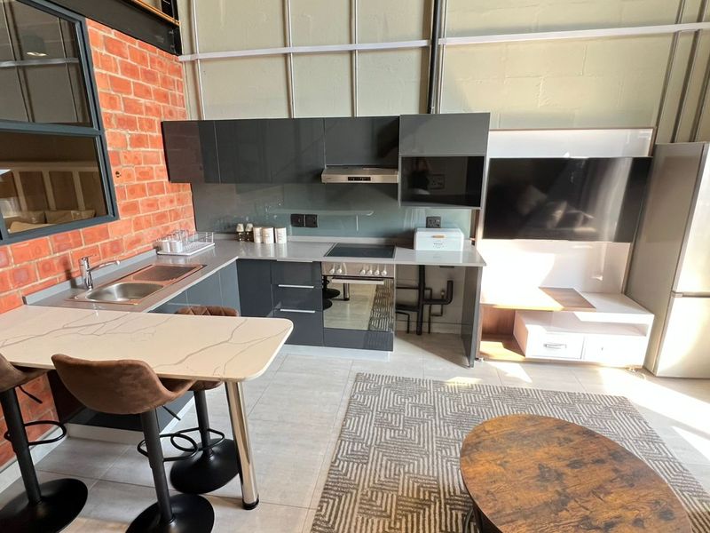 Modern open-plan kitchen and living area featuring sleek dark cabinetry, exposed brick accents, and integrated appliances in a contemporary industrial-style apartment.