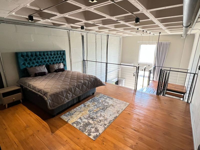 A spacious industrial-style mezzanine bedroom featuring exposed concrete waffle ceilings, timber flooring, and a metal balustrade overlooking the lower level.