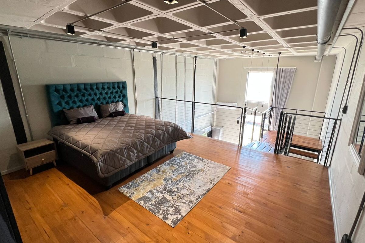 A spacious industrial-style mezzanine bedroom featuring exposed concrete waffle ceilings, timber flooring, and a metal balustrade overlooking the lower level.