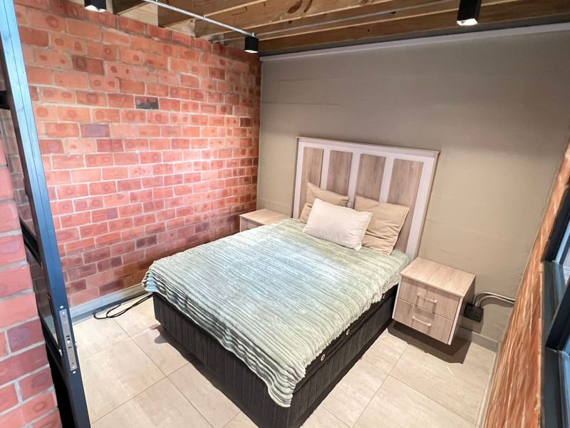 A modern bedroom featuring an exposed brick feature wall, industrial-style timber ceiling beams, track lighting, and a sleek steel-framed glass partition.