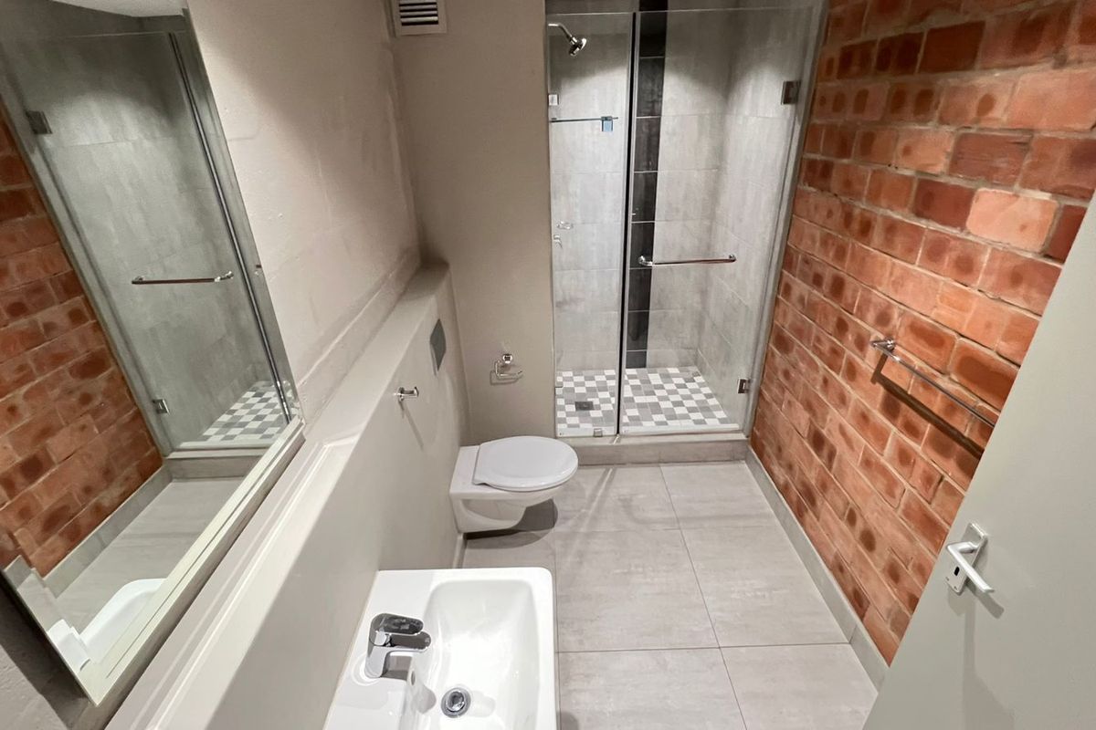 Modern bathroom featuring an exposed brick feature wall, glass shower enclosure, wall-mounted toilet, and contemporary washbasin with tiled flooring.
