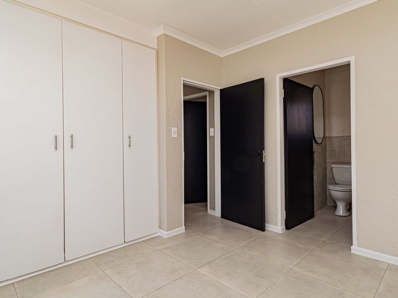 A clean, empty bedroom featuring neutral tiled flooring, built-in white cupboards, and direct access to an en-suite bathroom.
