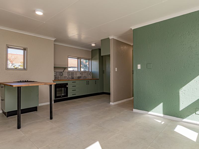 An open-plan kitchen and living area featuring modern green cabinetry, a built-in oven, patterned backsplash, and neutral tiled flooring throughout.