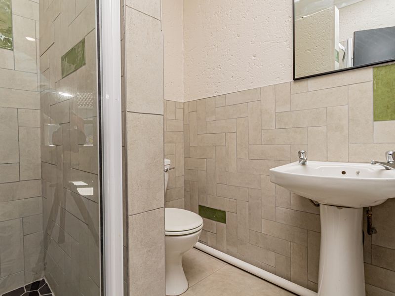 A functional bathroom featuring a pedestal basin, toilet, and a glass-enclosed shower with neutral wall tiling and exposed plumbing.