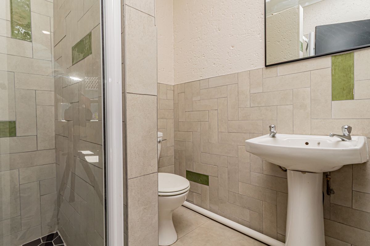 A functional bathroom featuring a pedestal basin, toilet, and a glass-enclosed shower with neutral wall tiling and exposed plumbing.