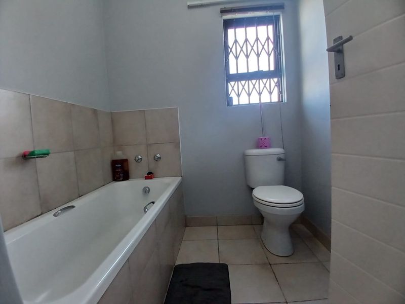 A functional bathroom featuring a standard bathtub, toilet, and neutral wall and floor tiling with a security-fitted window.