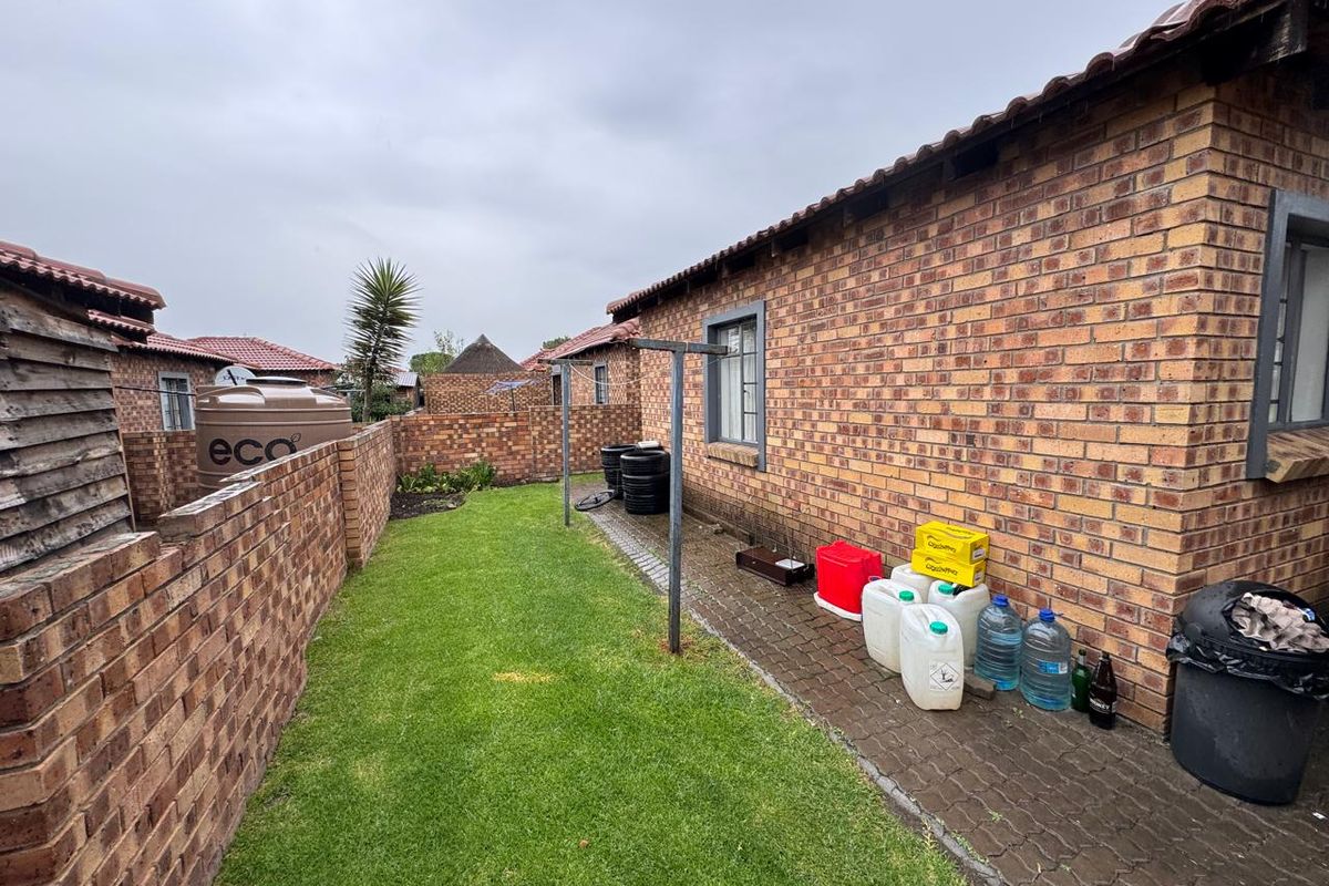 A rear exterior view of a townhouse featuring a face brick facade, a small lawn, a paved walkway, and a water storage tank.