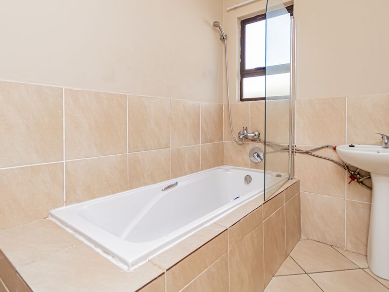 A functional bathroom featuring a built-in bathtub with a glass screen and a pedestal basin, finished with neutral ceramic wall and floor tiling.