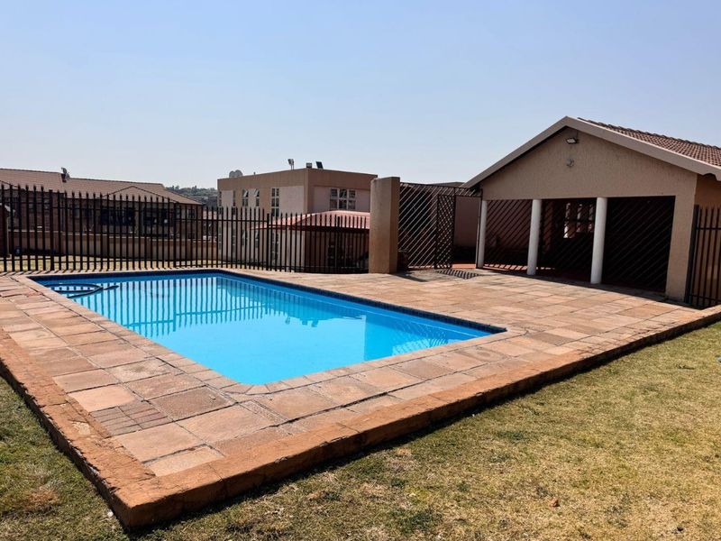 Outdoor pool area featuring a rectangular swimming pool, paved surround, and a covered patio area with structural columns, enclosed by metal perimeter fencing.